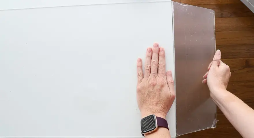 Hands guide a scoring tool along a white acrylic sheet, demonstrating how to cut acrylic sheet for a project.