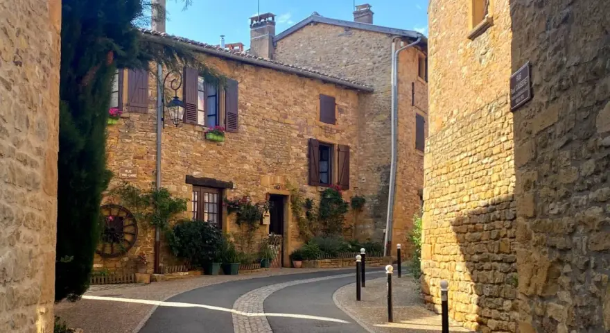 Village pittoresque aux maisons de pierre dorée, avec une ruelle sinueuse bordée de végétation luxuriante.