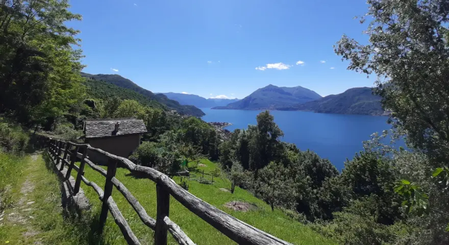 Sentiero del Viandante tappe: un casolare in pietra con tetto in ardesia e un recinto in legno rustico si affacciano su un lago blu intenso circondato da montagne verdi.