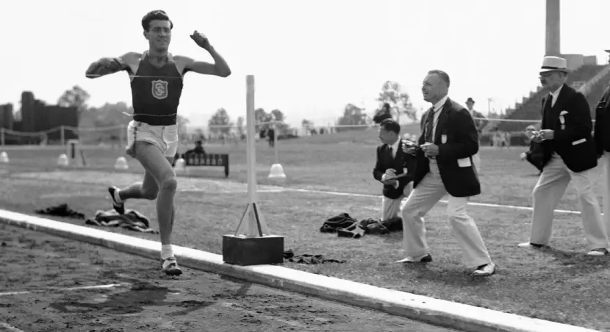 Louis Zamperini biegnie na bieżni, podczas gdy sędziowie obserwują zawody.