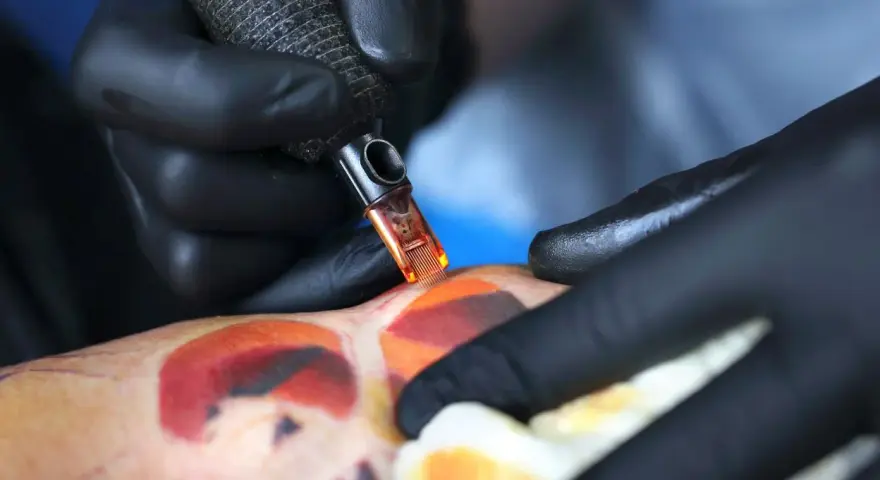Artysta w czarnych rękawiczkach tworzy kolorowy tatuaż. Pielęgnacja świeżego tatuażu jest kluczowa, a pytanie "ile razy dziennie myc tatuaz" jest ważne dla jego prawidłowego gojenia.
