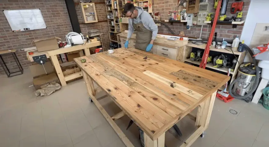 Uomo con grembiule lavora su un banco da lavoro con pallet, applicando finitura al legno. L'officina è piena di attrezzi e materiali.