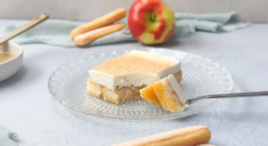 Ein Stück Apfel Schmand Kuchen mit Löffelbiskuits und einem Apfel im Hintergrund.