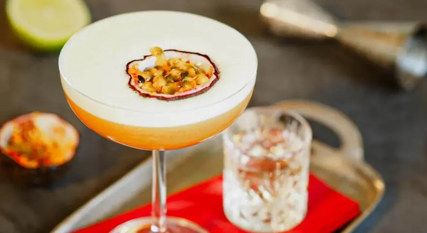 Orzeźwiający drink z martini, ozdobiony połówką marakui, obok kieliszka z wodą i połówki limonki.