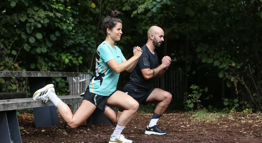Un couple fait des exercices de renfo course à pied en extérieur, avec des fentes et des squats.