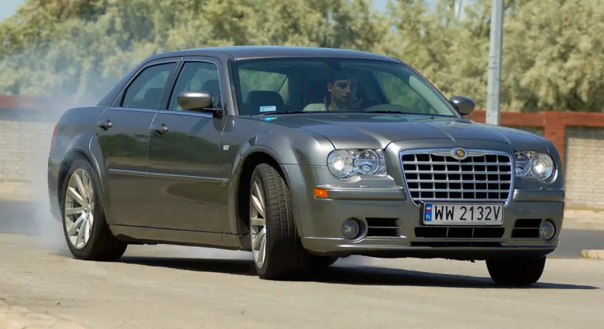 Szary Chrysler 300S z dymiącymi oponami, kierowca za kierownicą, na drodze.