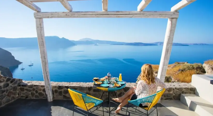 Mujer desayunando en balcón con vista al mar azul y montañas.