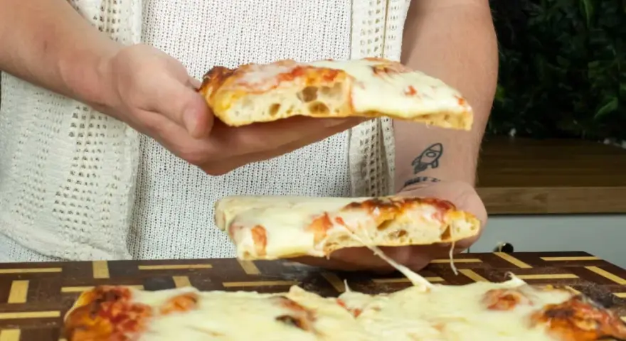 Due fette di pizza in teglia, con mozzarella filante e crosta dorata, vengono sollevate da mani.