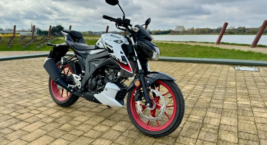 Biało-czarny Suzuki GSX S125 z czerwonymi felgami stoi na wybrukowanym placu nad jeziorem.