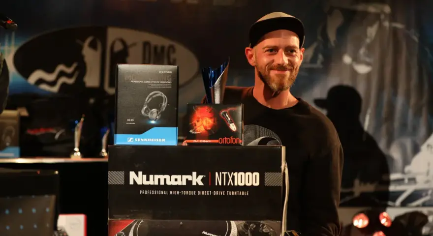 Un DJ souriant pose avec des prix et du matériel, dont des casques Sennheiser et un tourne-disque Numark. Une liste DJ internationale est visible.