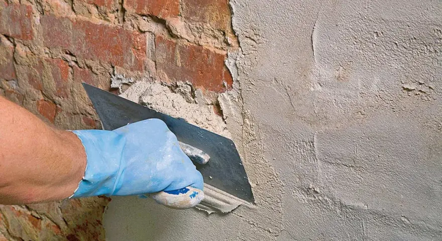 Hand mit blauer Handschuh glättet mit Kelle den **Sanierputz Keller** auf rotem Ziegelmauerwerk.