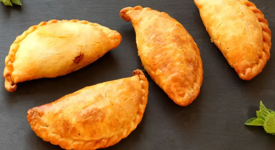 Cuatro empanadillas doradas y crujientes dispuestas sobre una superficie oscura, con hojas de menta a los lados.