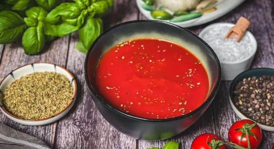 Schüssel mit leuchtend roter Pizza Sauce, umgeben von frischem Basilikum, Tomaten, Oregano, Salz und Pfeffer – wie in der Pizzeria.