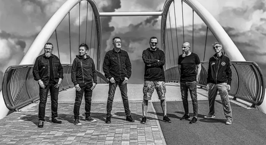 Sześciu mężczyzn, zespół Strachy na Lachy, pozuje na moście. Gotowi na koncerty, w rockowym stylu, pod burzliwym niebem.
