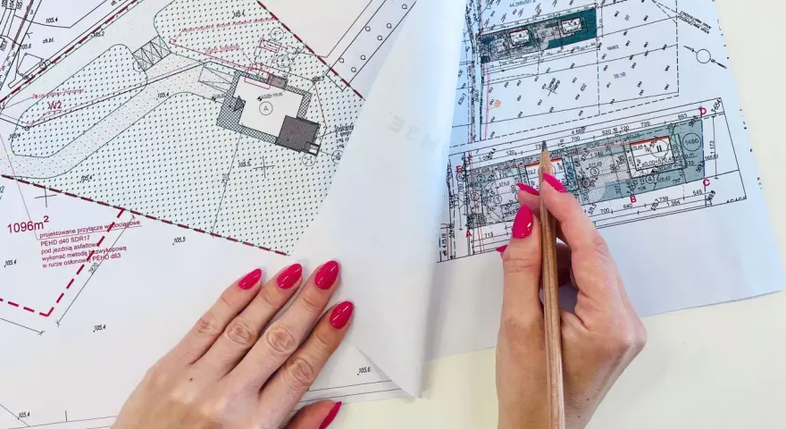 Dłoń z różowym lakierem na paznokciach trzymająca ołówek nad mapą projektową.