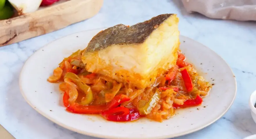 Un delicioso bacalao encebollado reposa sobre una cama de pimientos y cebolla.