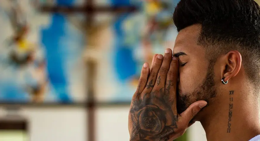 A man with tattoos prays before a crucifix, contemplating if is vaping a sin.