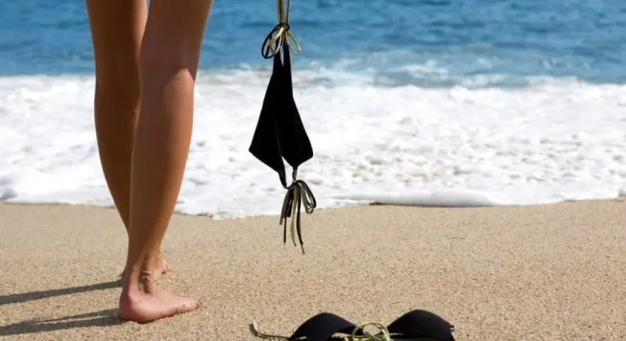 Nogi na piaszczystej plaży, obok czarny kostium kąpielowy. Fale obmywają brzeg. Idealne miejsce na naturystyczną plażę.