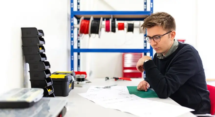 Młody mężczyzna w okularach analizuje dokumentację techniczną. Na stole leżą narzędzia, a w tle widać półkę z przewodami, co sugeruje pracę nad projektem z dziedziny plan bioz.