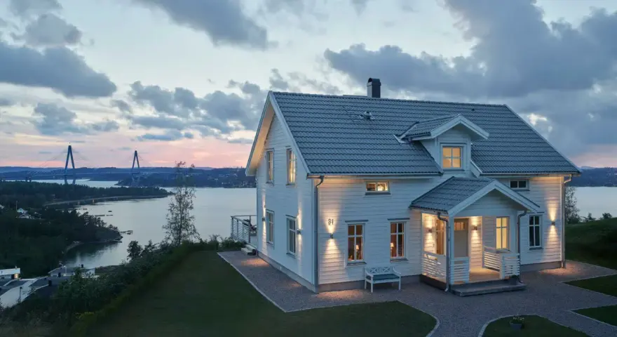 Vackert vitt hus med upplysta fönster och entré, perfekt hus entre inspiration. Vacker vy över vatten och bro i skymningen.