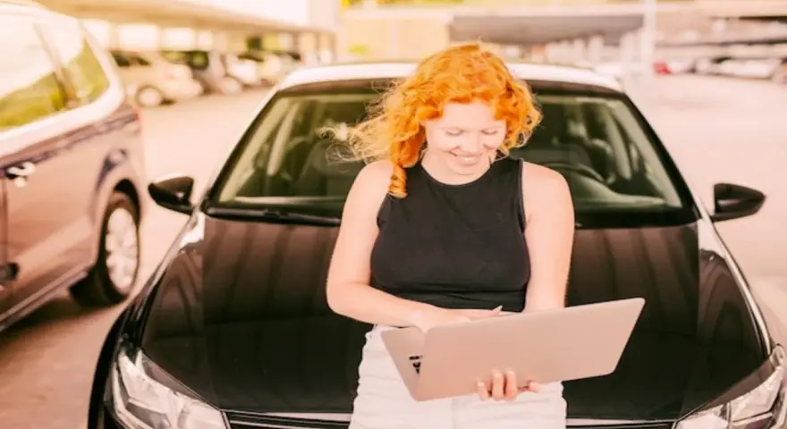Kobieta z rudymi włosami uśmiecha się, trzymając laptopa. Za nią czarny samochód, być może przygotowania do rejestracji samochodu z Niemiec.