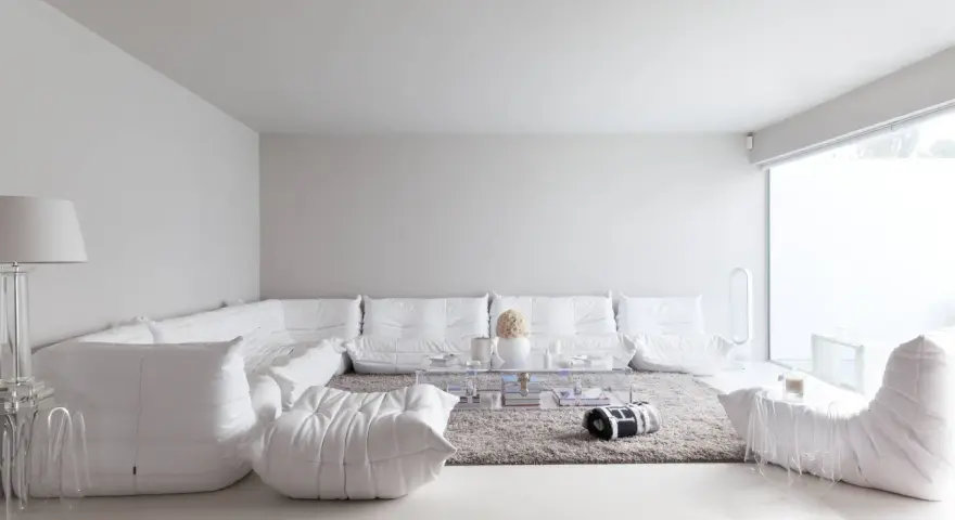 Soggiorno minimalista con divano componibile bianco, tappeto grigio e tavolino trasparente. Le tonalità di bianco per pareti creano un'atmosfera eterea.