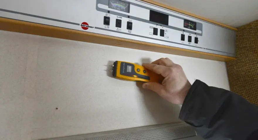 Hand håller fuktmätare mot vägg för fukttest protokoll.
