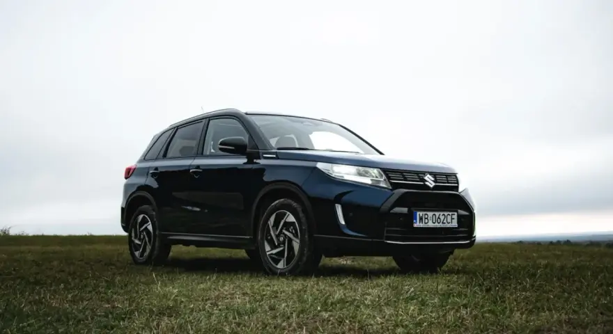 Ciemnoniebieski Suzuki Vitara Hybrid na tle zachmurzonego nieba, zaparkowany na trawiastym wzgórzu.