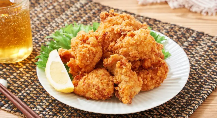 Chrupiące karaage chicken na talerzu, ozdobione plasterkiem cytryny i liśćmi sałaty. Obok szklanka złocistego napoju i pałeczki.