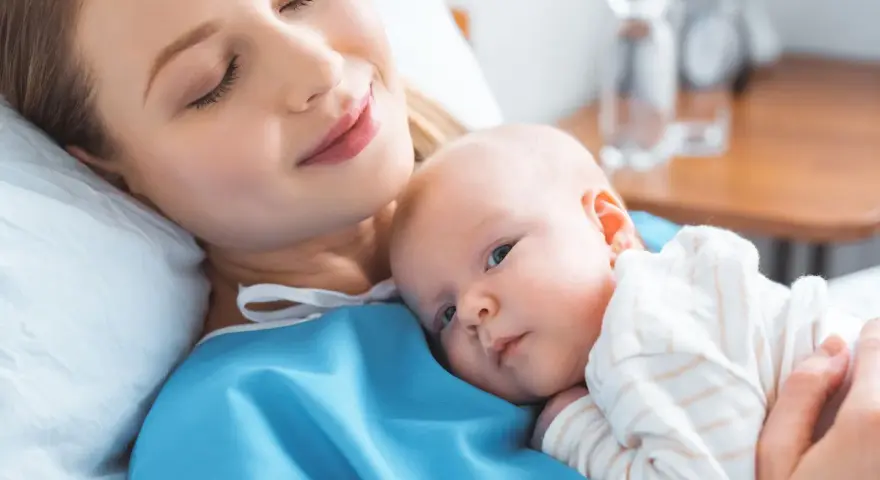 Uśmiechnięta mama tuli noworodka. To chwila spokoju po szczepieniach noworodka, budująca więź i zapewniająca zdrowie maluszka.