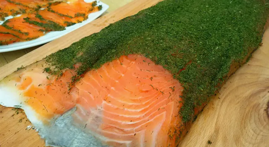 Un gran filete de salmón marinado casero cubierto de eneldo fresco. En el fondo, finas lonchas de salmón ya preparadas.