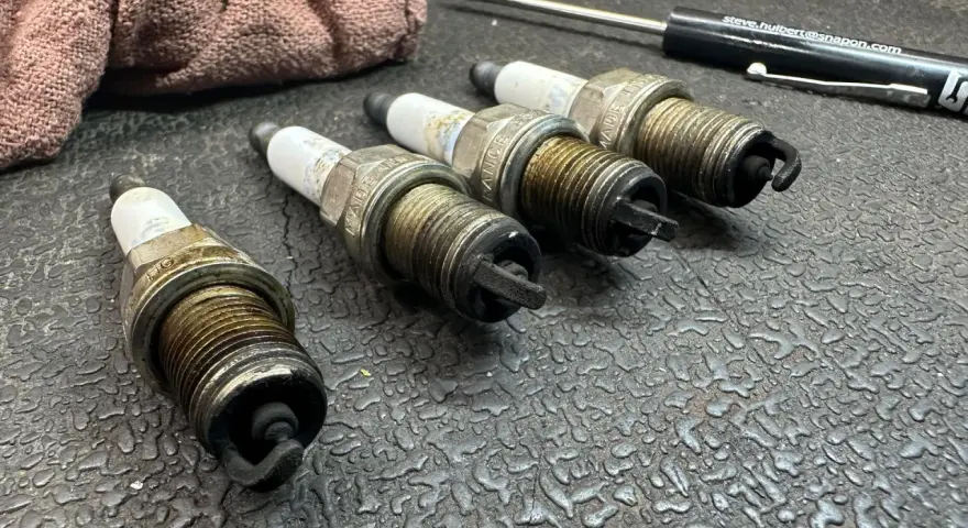 Four used spark plugs lie on a textured surface next to a rag and a screwdriver. This image might make you wonder how many spark plugs a car has.