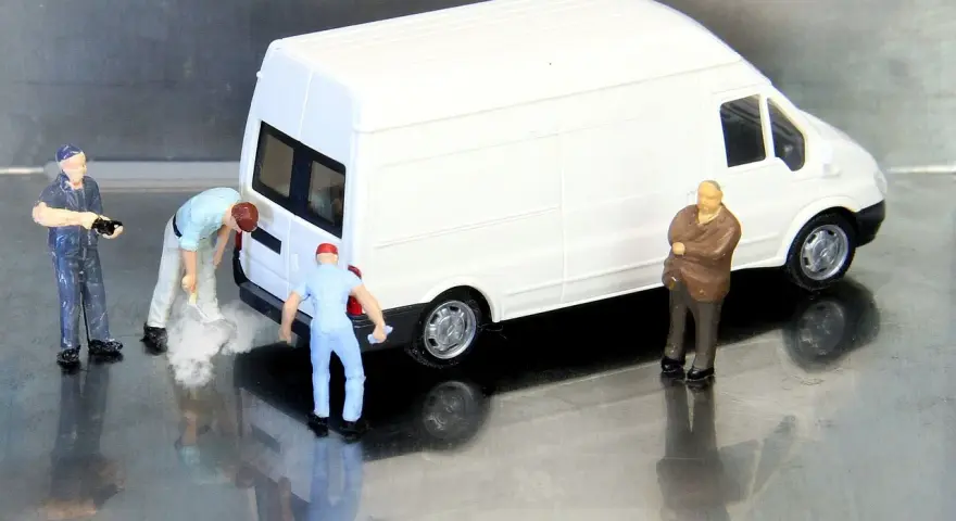 Miniaturowi pracownicy zajmują się białym busem budowlanym. Jeden z nich czyści tył pojazdu, inny trzyma narzędzie, a trzeci obserwuje.