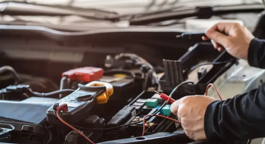 Mechanik sprawdza instalację elektryczną samochodu. Na zdjęciu widać multimetr i ręce trzymające sondy. Stojan z bezpiecznikami jest widoczny w tle.