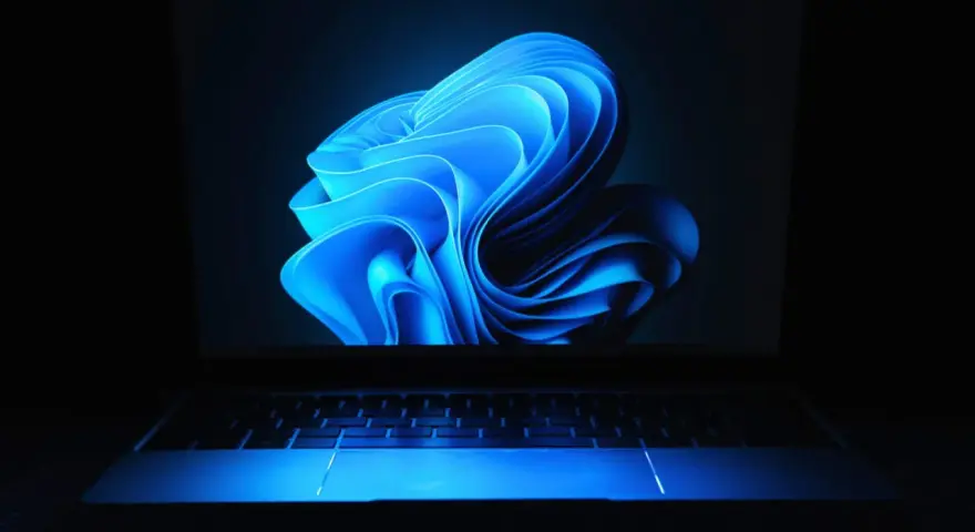 Niebieski, falujący kształt na ekranie laptopa z podświetlaną klawiaturą, przypominający logo Windows 11 24H2.