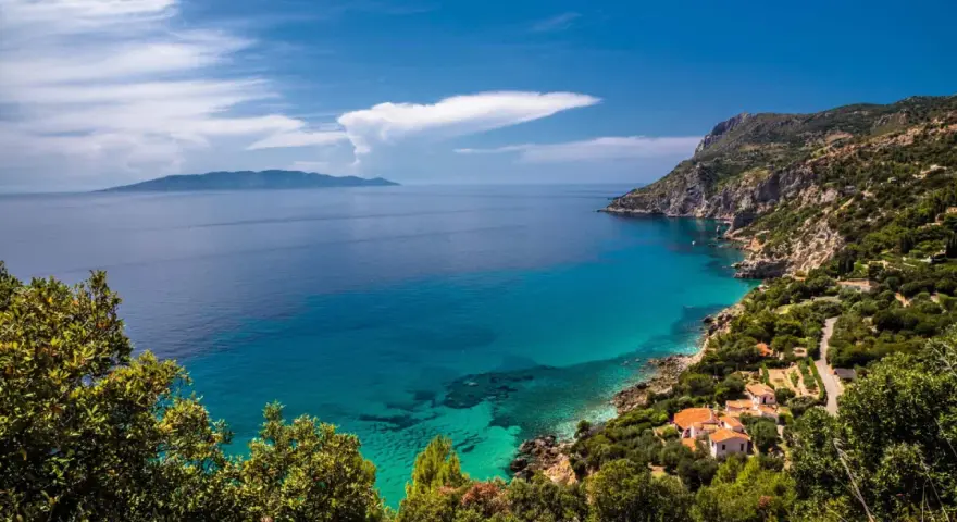 Turquoise water laps a rocky Tuscan coast with villas nestled among trees, under a blue sky with clouds.