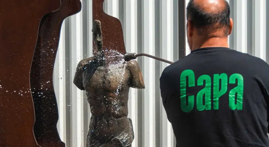 Hombre con camiseta negra "Capa" limpia una escultura de bronce con agua a presión.