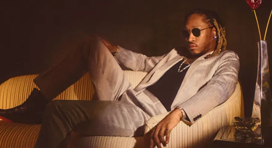Ein Hip-Hop-Künstler in einem schicken Anzug entspannt sich auf einem Sofa, trägt Sonnenbrille und Schmuck.