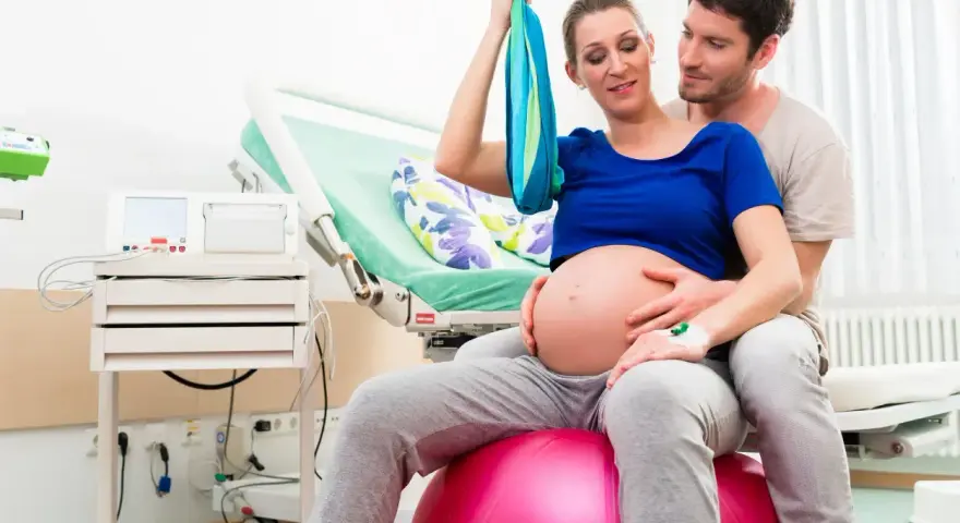 Para z niecierpliwością oczekuje porodu. Kobieta siedzi na piłce porodowej, wspierając się na chuście. Mężczyzna obejmuje ją czule. To może być ich pierwszy poród, ale przygotowują się na VBAC.