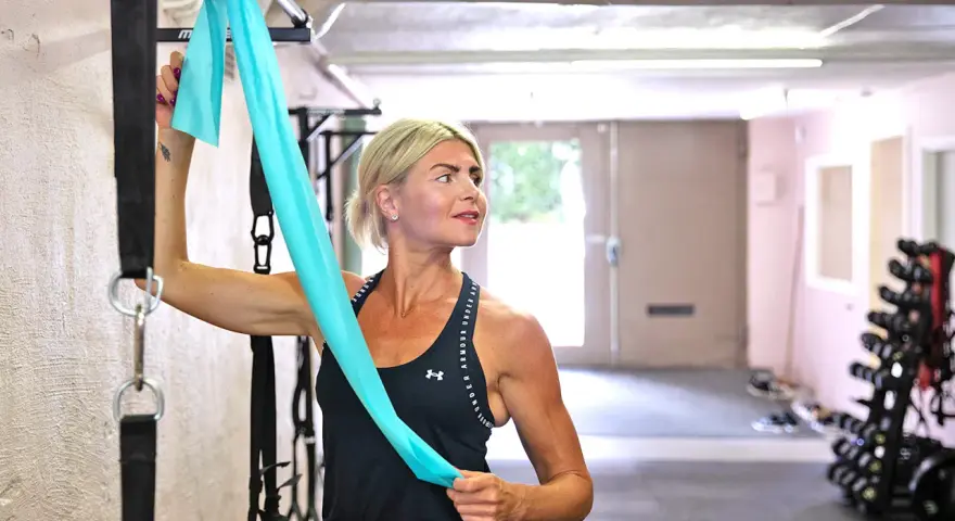 Kvinna med blont hår stretchar axlar med ett turkost träningsband i ett gym.