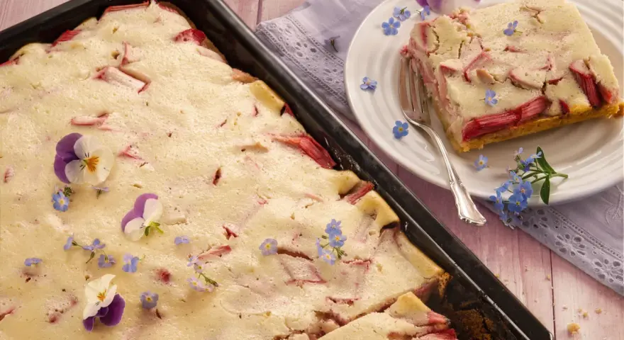 Saftiger Rhabarberkuchen mit Quark, dekoriert mit Veilchen und Vergissmeinnicht. Ein Stück ist auf einem Teller angerichtet.