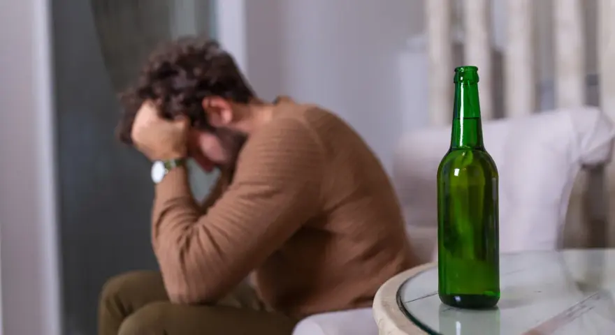 Mężczyzna siedzi z głową w dłoniach, obok stoi zielona butelka. Czuje duszności po alkoholu.