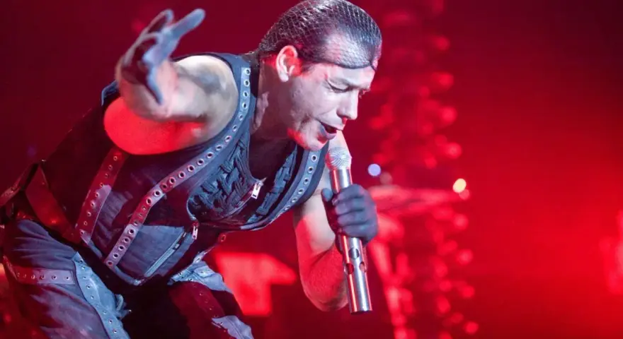 Till Lindemann, chanteur du groupe metal allemand Rammstein, en pleine performance sur scène, micro à la main, sous un éclairage rouge intense.