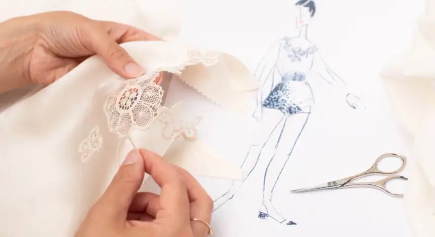 Main d'une couturière réalisant un assemblage couture délicat sur un tissu de soie, avec un croquis de mode et des ciseaux à proximité.