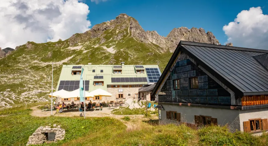 Die Buchel Alpe, eine Berghütte mit Solaranlagen, liegt vor einer beeindruckenden Bergkulisse. Gäste sitzen draußen unter Sonnenschirmen.