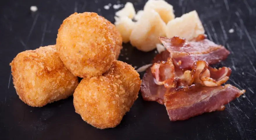 Croquetas doradas y crujientes con trozos de bacon y queso sobre fondo oscuro.