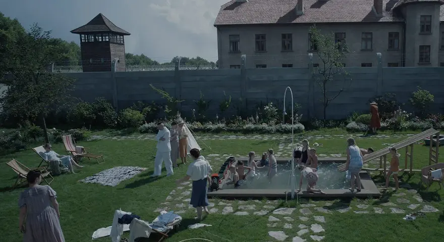 Scena da film strani: bambini giocano in piscina, adulti sorvegliano, mentre sullo sfondo incombono torri di guardia e un edificio imponente.