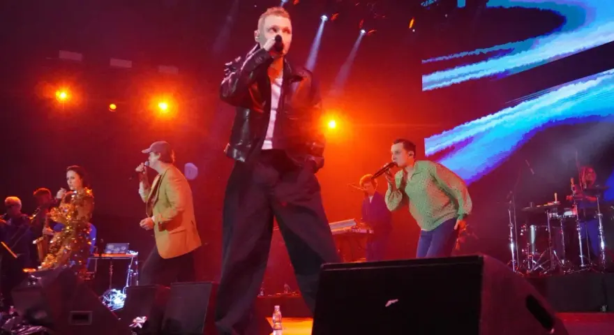 Męskie granie hymny na żywo. Wokalista w skórzanej kurtce i szerokich spodniach śpiewa do mikrofonu. Na scenie inni artyści i muzycy.