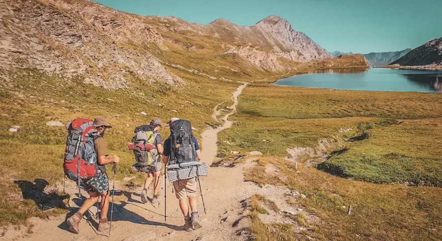 Trois randonneurs équipés pour le tour du Queyras traversent un sentier de montagne, un lac bleu et des sommets rocheux en toile de fond.