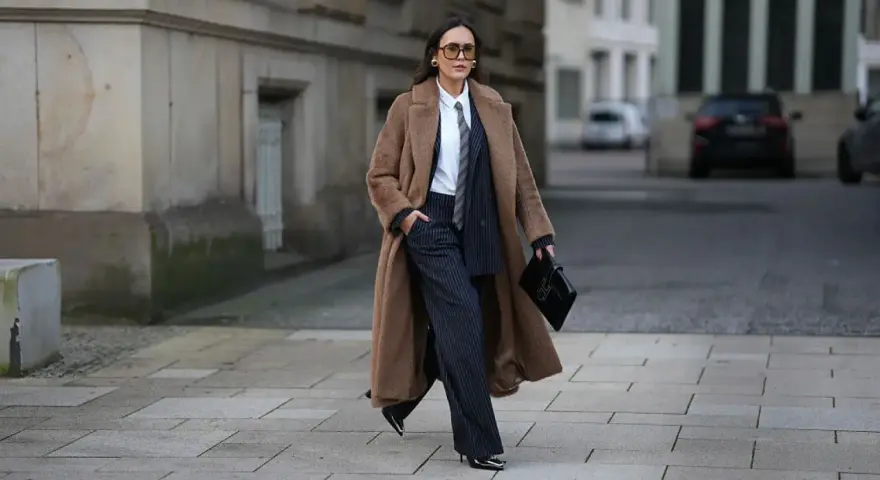 Chica tomboy con abrigo largo marrón, traje de raya diplomática y bolso negro.